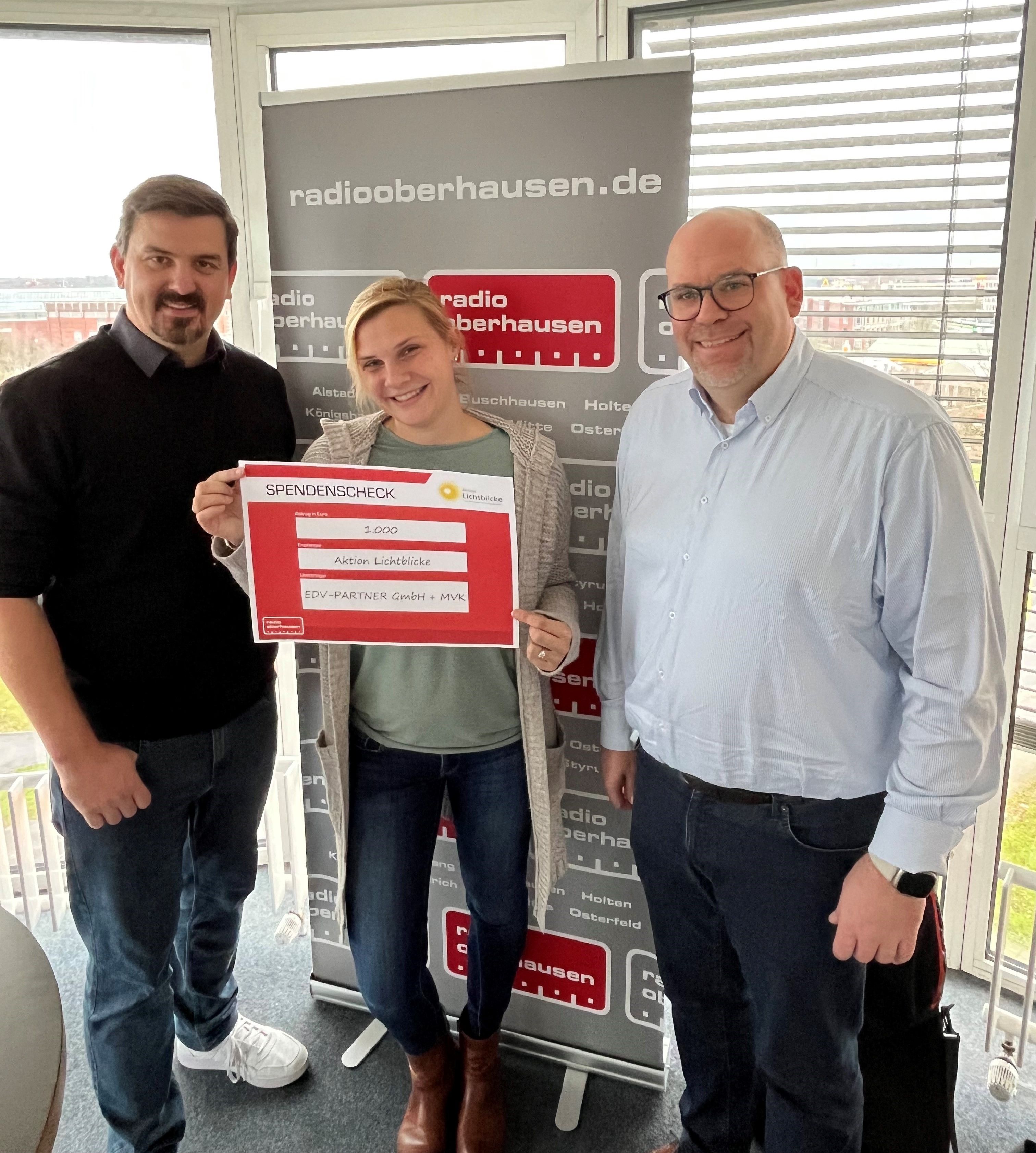 Bastian Schumacher, Geschäftsführer von EDV-PARTNER, Veit Kuhl, Geschäftsführer von der MVK GmbH und die Reporterin von Radio Oberhausen halten gemeinsam einen Spendencheck über 1000 €, adressiert an die Aktion Lichtblicke
